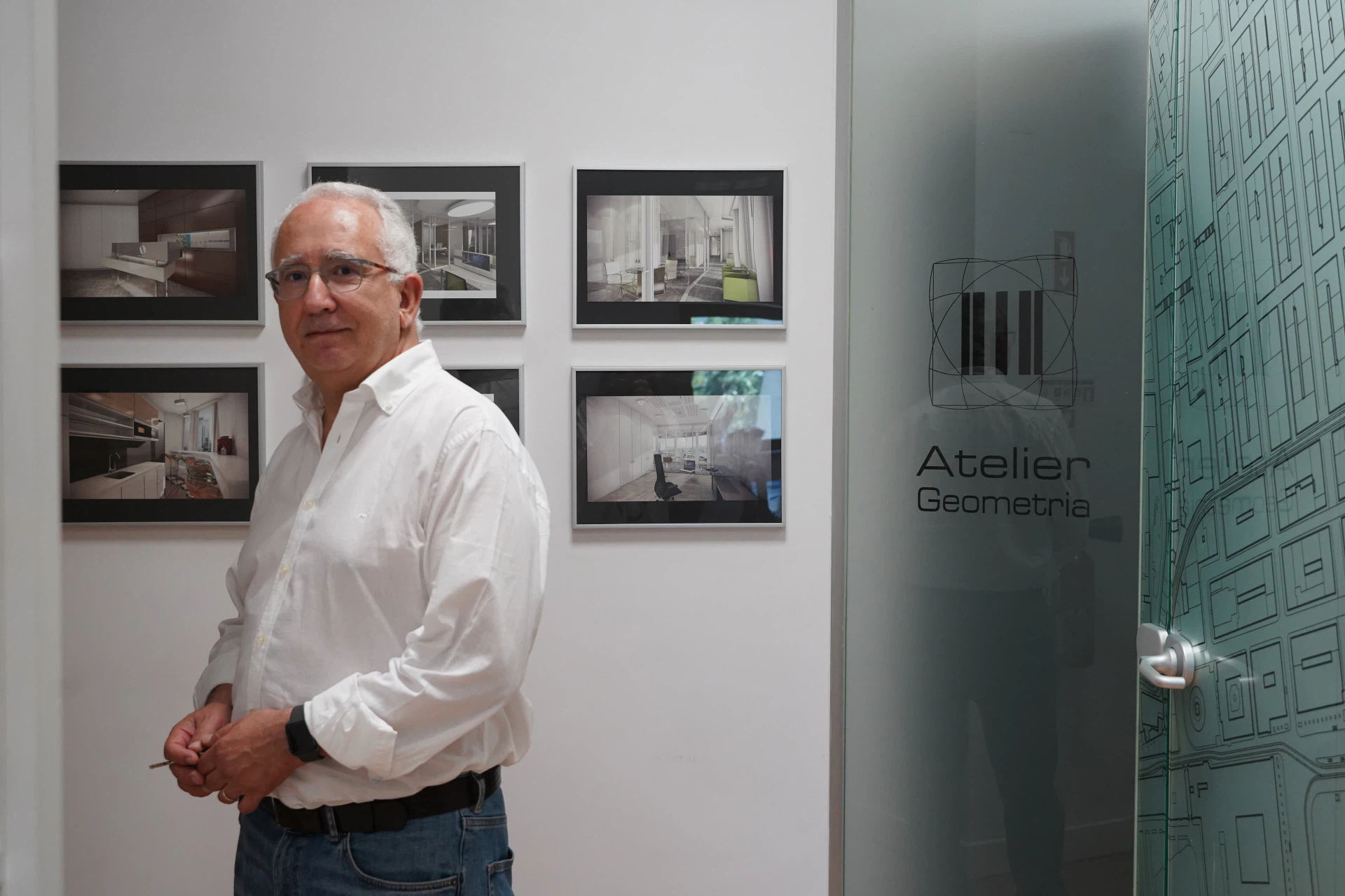 Arquiteto no atelier, junto à parede com imagens de projetos emolduradas e à porta de vidro gravada com a marca Atelier Geometria