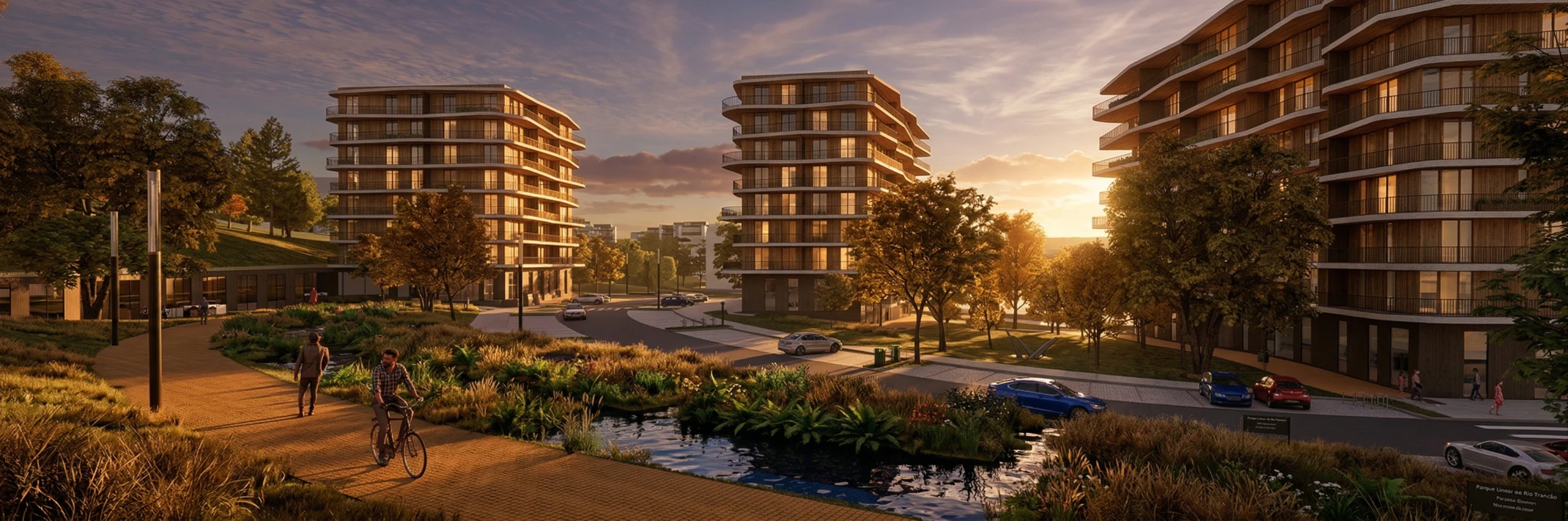 Os edifícios de habitação vistos do parque ao fim da tarde: o percurso em madeira por onde passam um peão e um ciclista, a linha de água com vegetação ripícola e, ao fundo à direita, o piso térreo comercial sobre a rua.